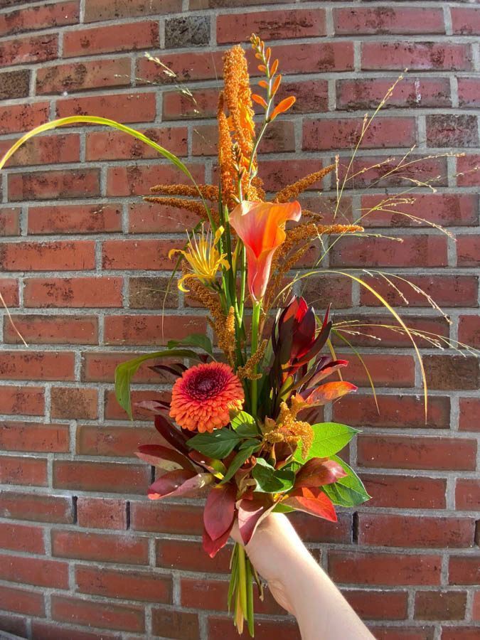 Ein handgehaltener Blumenstrauß mit orangefarbenen, roten und grünen Blüten vor einer Backsteinmauer.