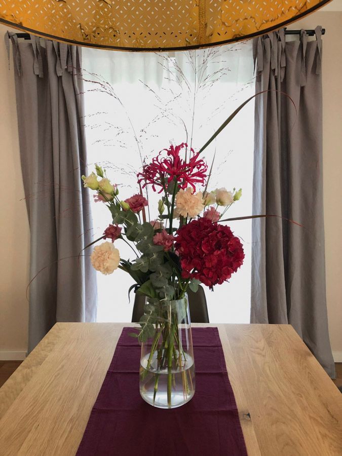Eine Glasvase mit gemischten Blumen auf einem Holztisch mit einem violetten Tischläufer, vor grauen Vorhängen.