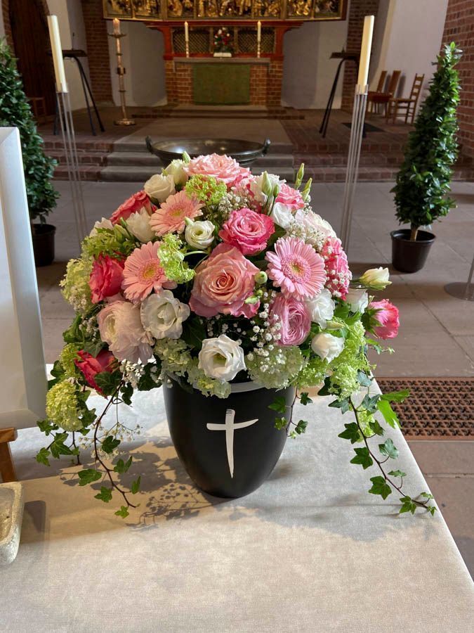 Ein rosa-weißes Blumenarrangement in einer schwarzen Vase mit einem Kreuz, aufgestellt in einem Kirchenraum.