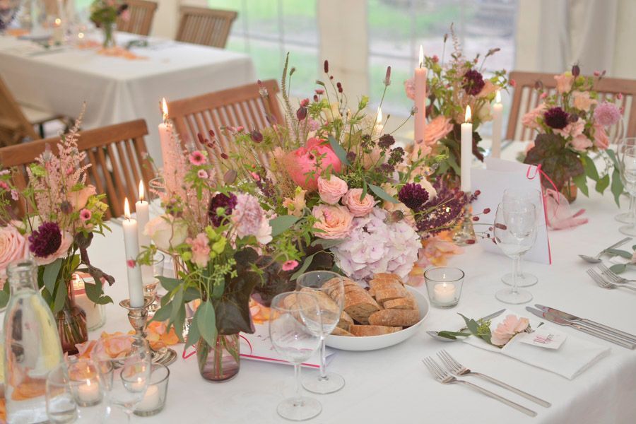 Elegante Hochzeitstafel mit rosa Blumengestecken, Kerzen, Weingläsern und Brot auf weißen Tischdecken.