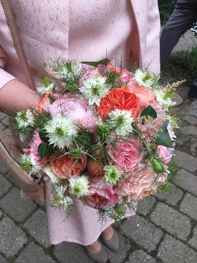 Eine Person hält einen Strauß rosa, korallenroter und weißer Blumen auf einem Kopfsteinpflasterweg