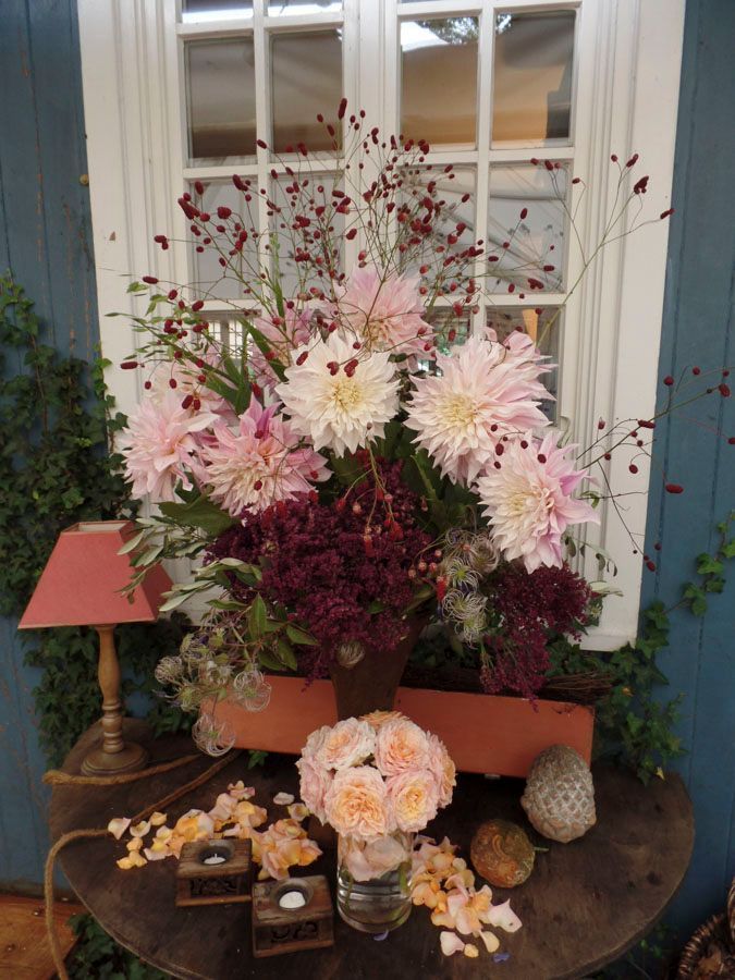 Ein Blumenarrangement mit hellrosa und weißen Blüten vor einem Fenster, auf einem Tisch mit Kerzen und kleiner Dekoration.