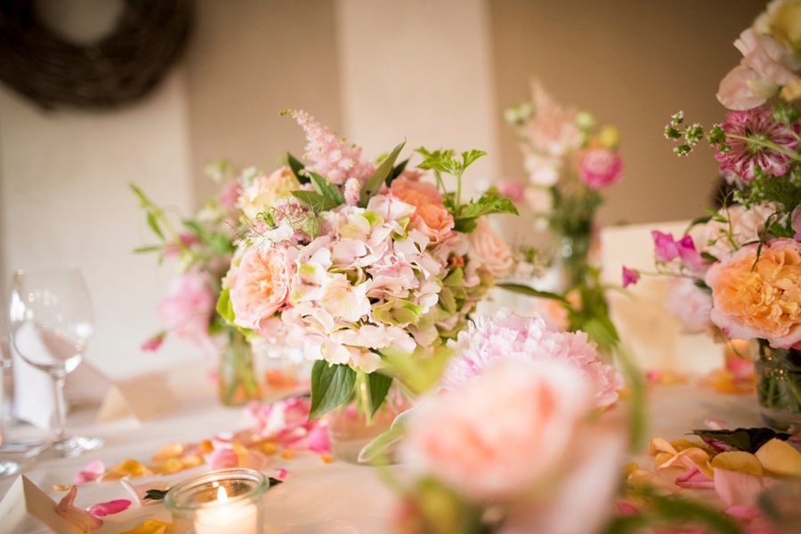 Pastellfarbenes Blumen-Tischgesteck mit rosa und pfirsichfarbenen Blüten, Kerzen und Gläsern auf einem dekorierten Tisch