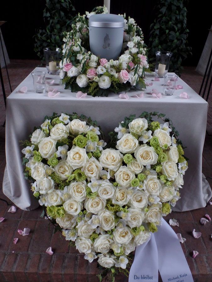 Ein herzförmiges Gesteck aus weißen Rosen unter einer grauen Urne auf einem Gedenktisch