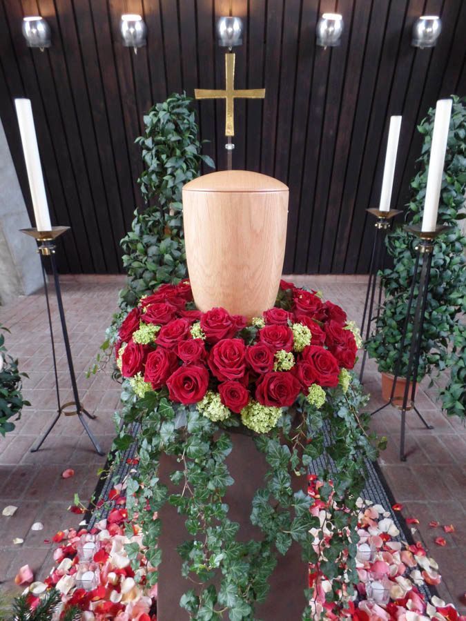 Eine hölzerne Urne mit einer roten Rose in der Mitte, umgeben von Grünzeug, brennenden Kerzen und einem Kreuz in einer Gedenkstätte.