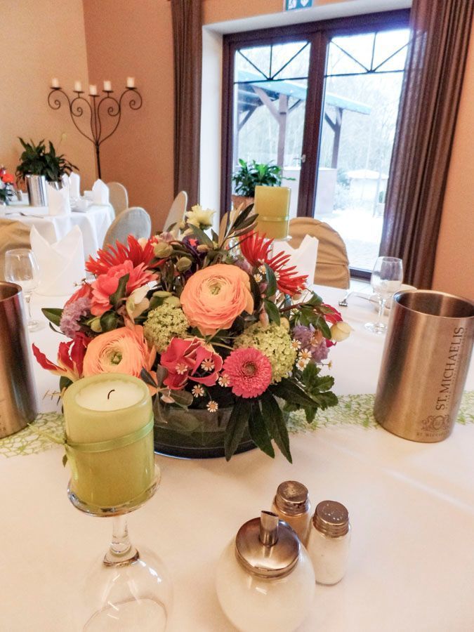 Ein Blumenarrangement als Tischmitte auf einem weißen Tisch mit Kerzen, Salz- und Pfefferstreuern und dem Blick auf das Esszimmer mit Fenster im Hintergrund.