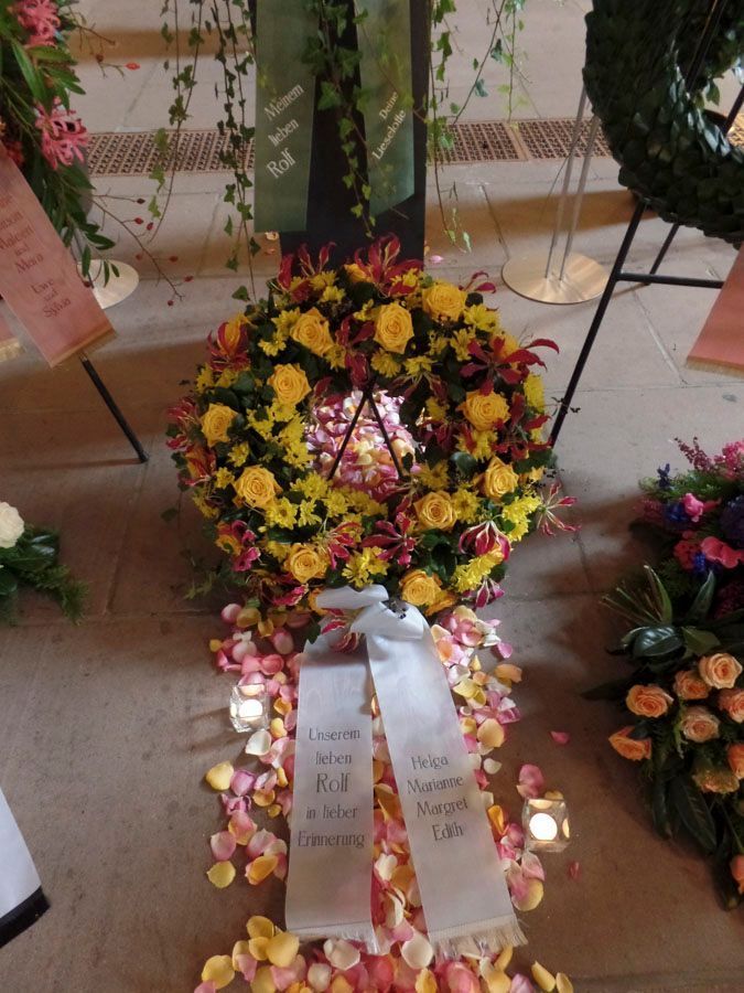 Ein Blumenkranz mit weißen Bändern und Kerzen, der im Innenbereich zwischen anderen Gedenkgestecken aufgestellt ist.