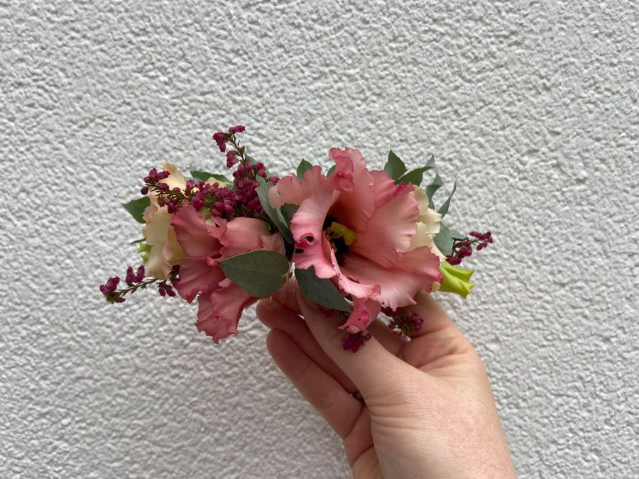 Eine Hand hält einen kleinen rosa-grünen Blumenstrauß vor einer strukturierten weißen Wand.