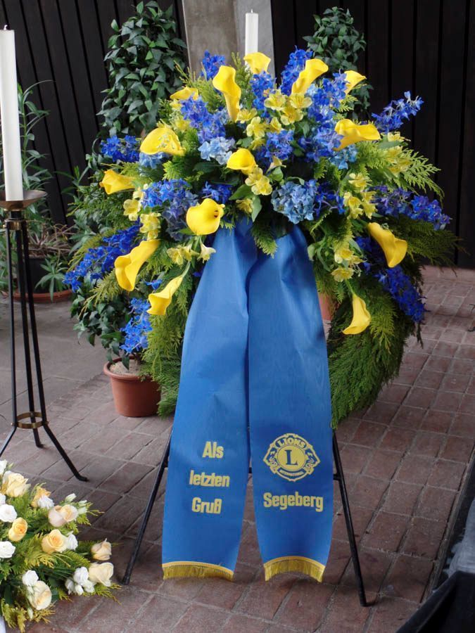 Blauer und gelber Blumenkranz auf einem Ständer mit einem Band, auf dem „Als letzten Gruß“ und „Segeberg“ steht