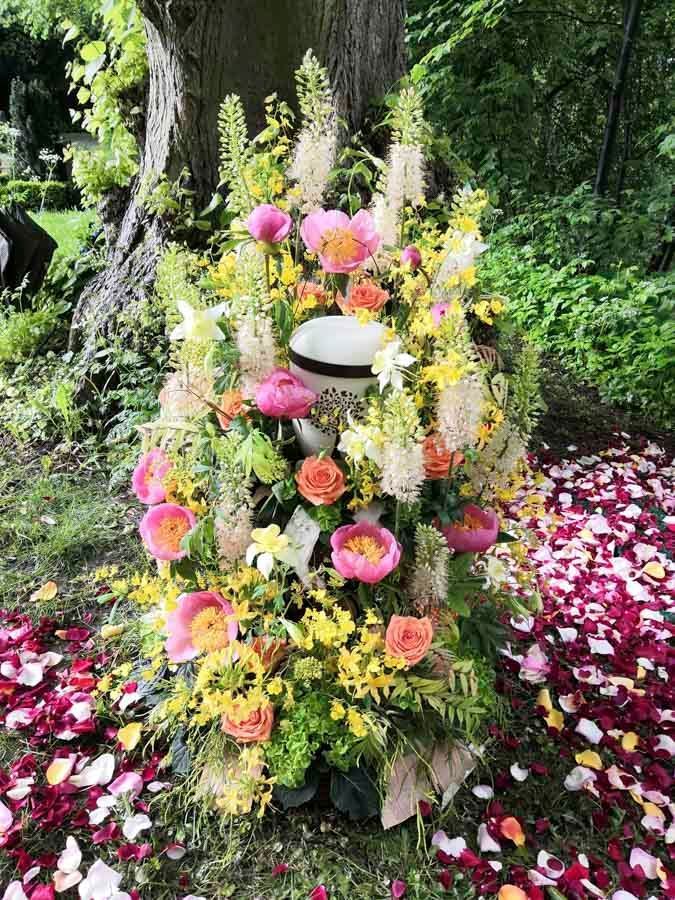 Blumengestecke als Gedenkstätte mit rosa, gelben und weißen Blüten am Fuße eines Baumes, Blütenblätter auf dem Boden verstreut.