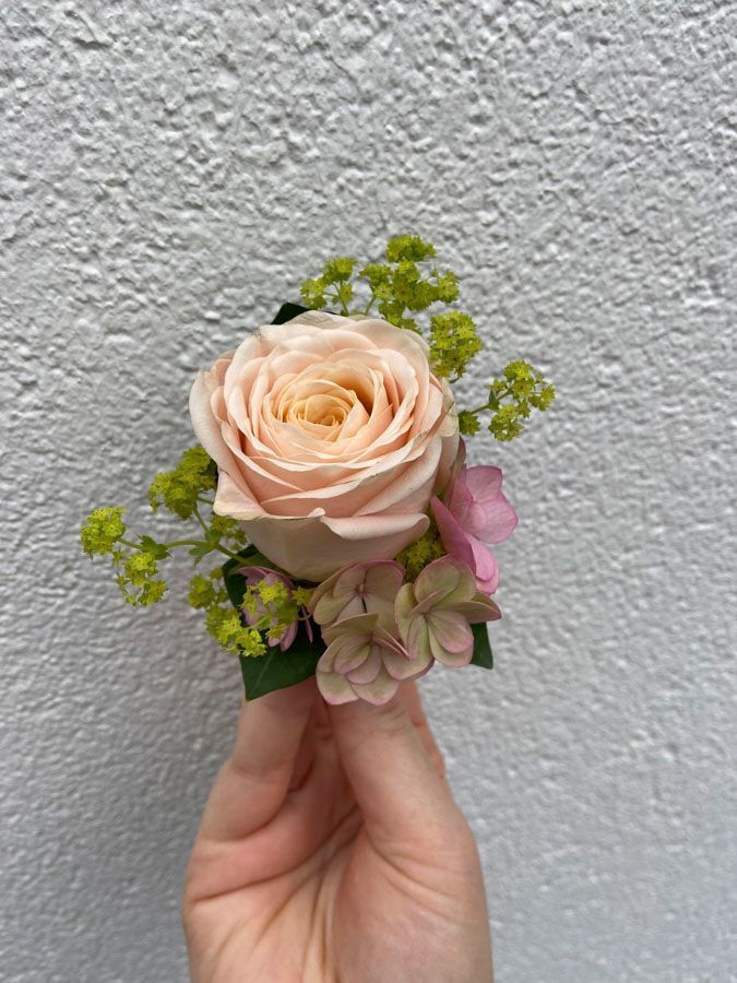 Eine Hand hält ein kleines pastellfarbenes Blumenarrangement mit einer pfirsichfarbenen Rose und grünem Füllmaterial vor einer grauen Wand.