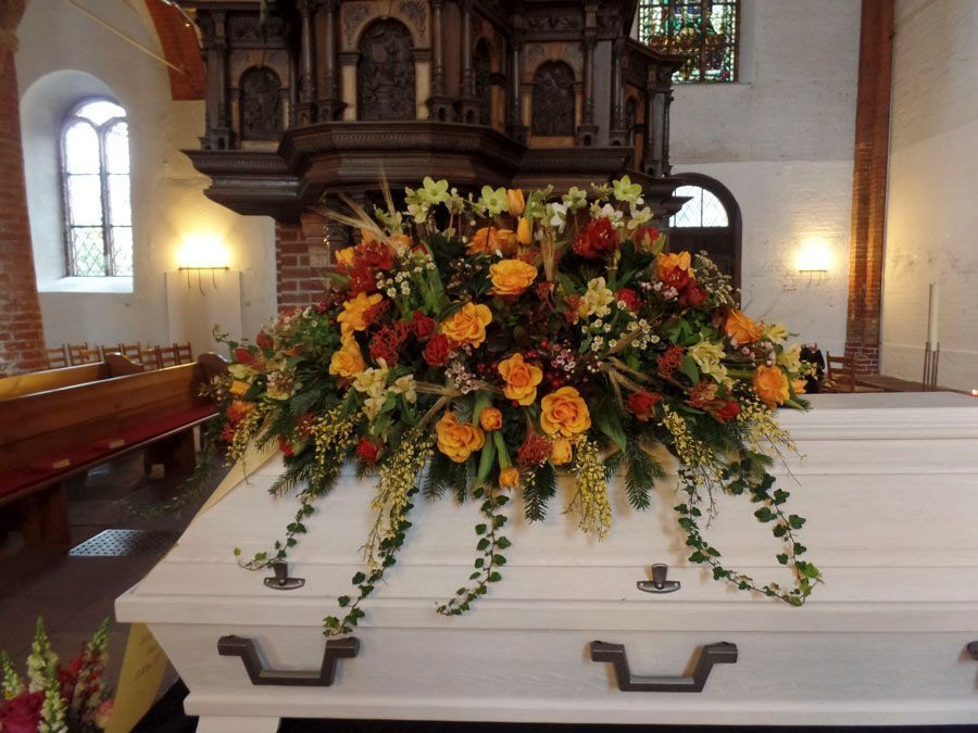 Blumenarrangement aus orangefarbenen, gelben und weißen Blumen auf einem weißen Sarg in einer Kirche