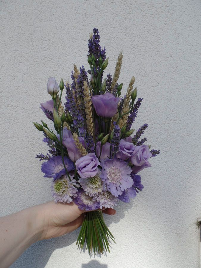 Ein handgehaltener Strauß aus lila Wildblumen und Lavendelgrün vor einer weißen Wand