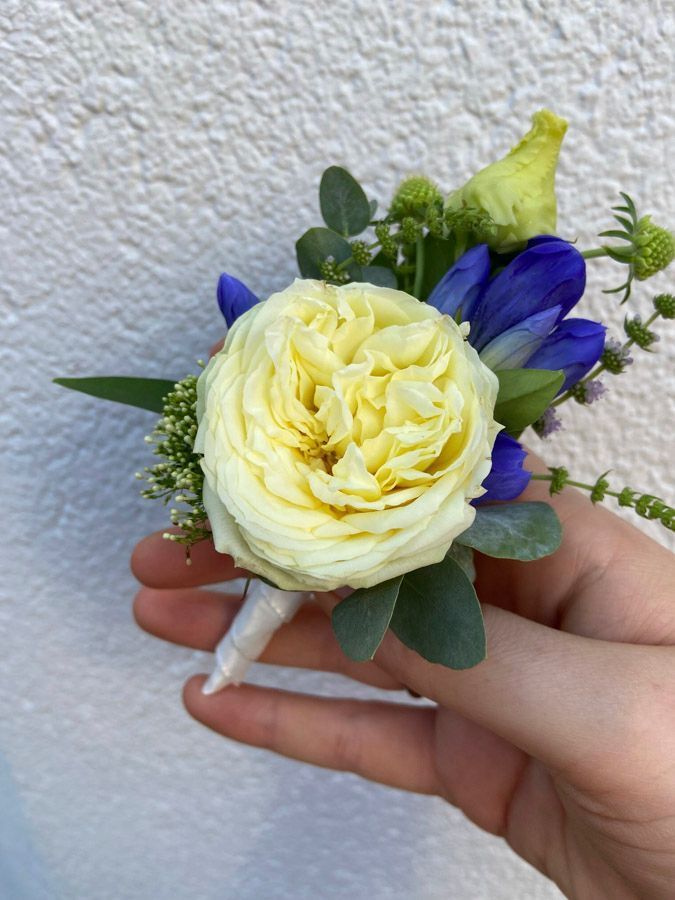 Eine Hand hält einen kleinen Strauß mit einer cremefarbenen Rose, lila Blüten, Grünzeug und einem umwickelten Stiel.