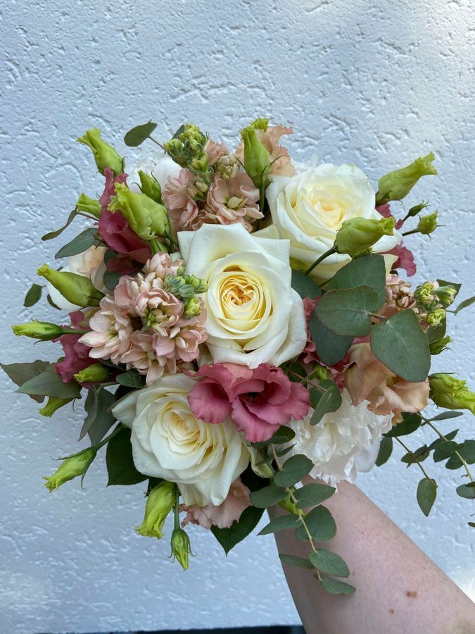 Ein handgehaltener Strauß aus weißen Rosen, rosa Blüten und Grün vor einer hellblauen Wand