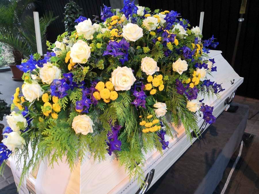 Blumengesteck für den Sarg mit weißen Rosen, gelben Blüten und violetten Blumen auf einem weißen Sarg