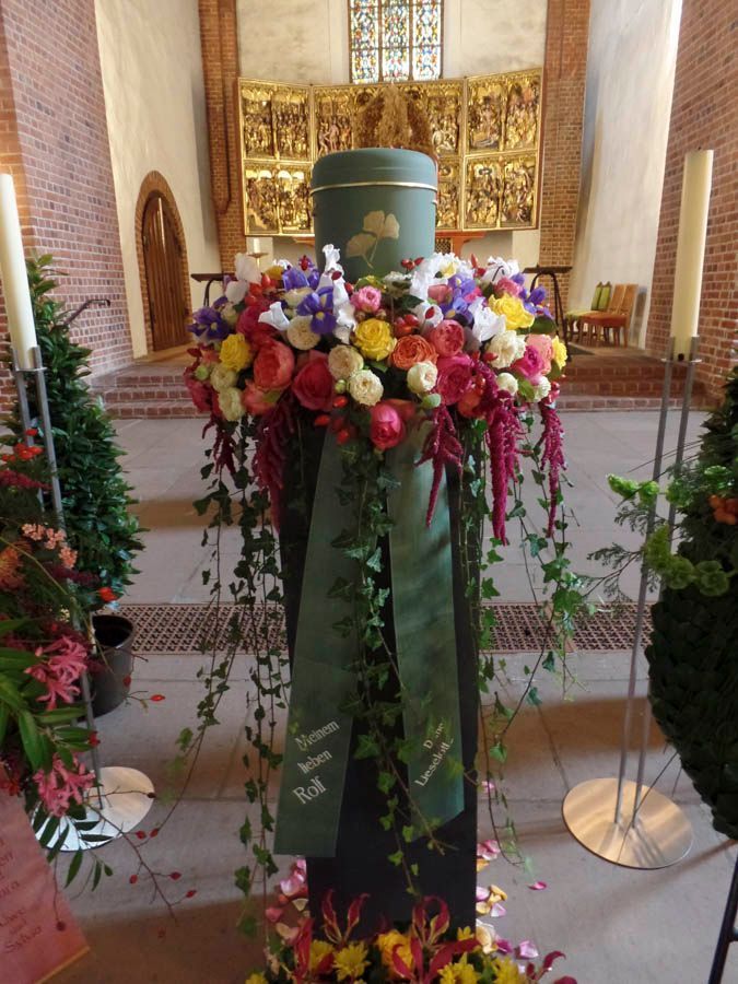 Ein mit Blumen bedeckter Gedenkplatz im Kirchenschiff, in der Nähe bunte Blumensträuße und Kerzen.