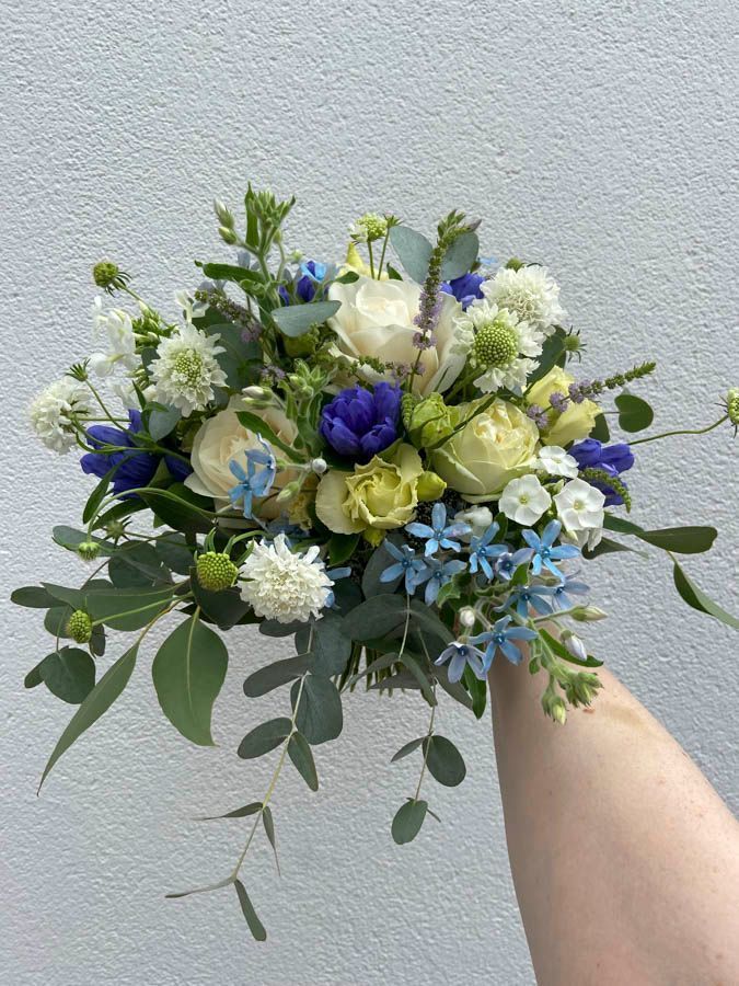 Ein handgehaltener Blumenstrauß aus weißen, blauen und grünen Blumen vor einer hellen Wand