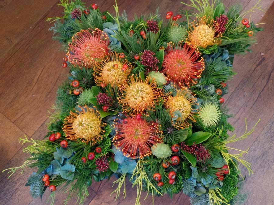 Quadratisches Blumenarrangement mit roten, gelben und grünen stacheligen Blüten und kiefernartigem Grün auf einem Holzboden