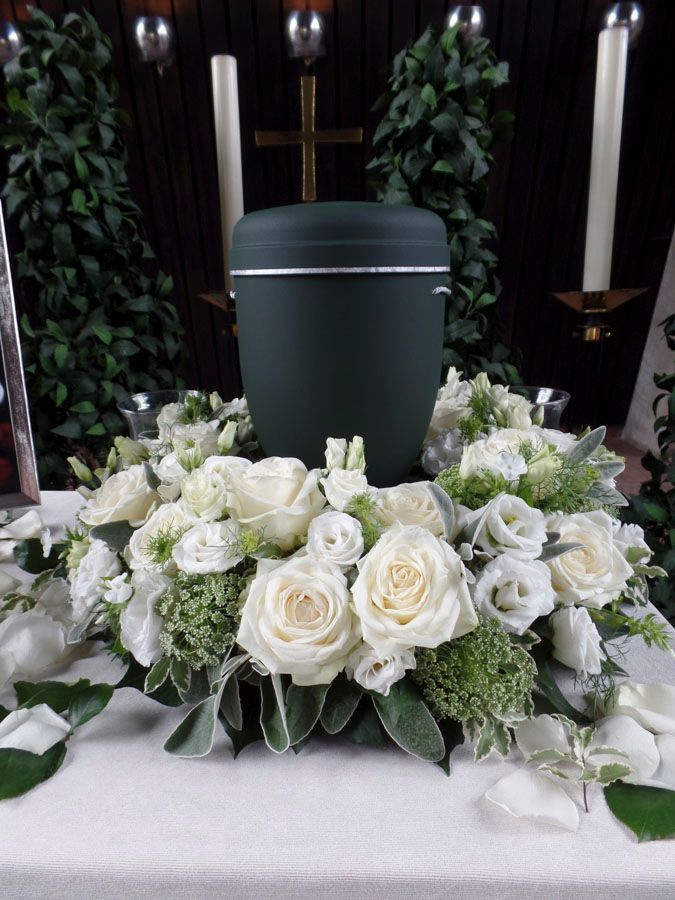 Dunkelgrüne Urne, umgeben von weißen Rosen und Grünpflanzen, vor einem weißen Stoffhintergrund