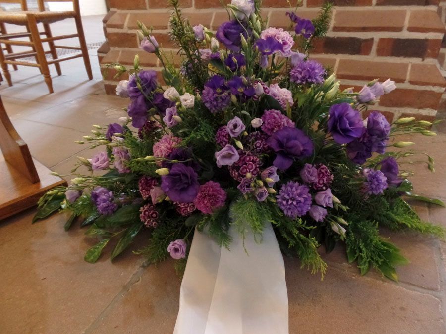 Lila Blumenarrangement mit weißem Band auf einem Fliesenboden neben einer Backsteinmauer
