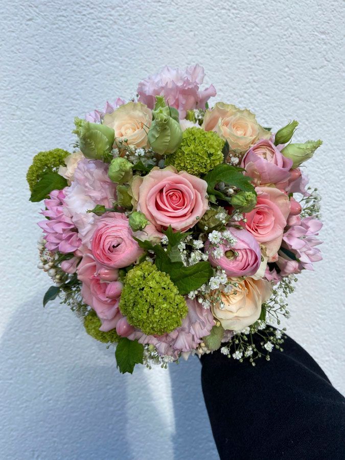 Ein handgehaltener Blumenstrauß aus rosa, cremefarbenen und grünen Blumen vor einer hellblauen Wand