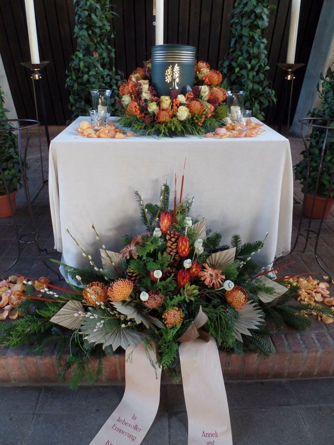 Blumengeschmückter Traueraltar mit weißem Tuch, orangefarbenen und weißen Gestecken und Gedenkbändern auf den Stufen