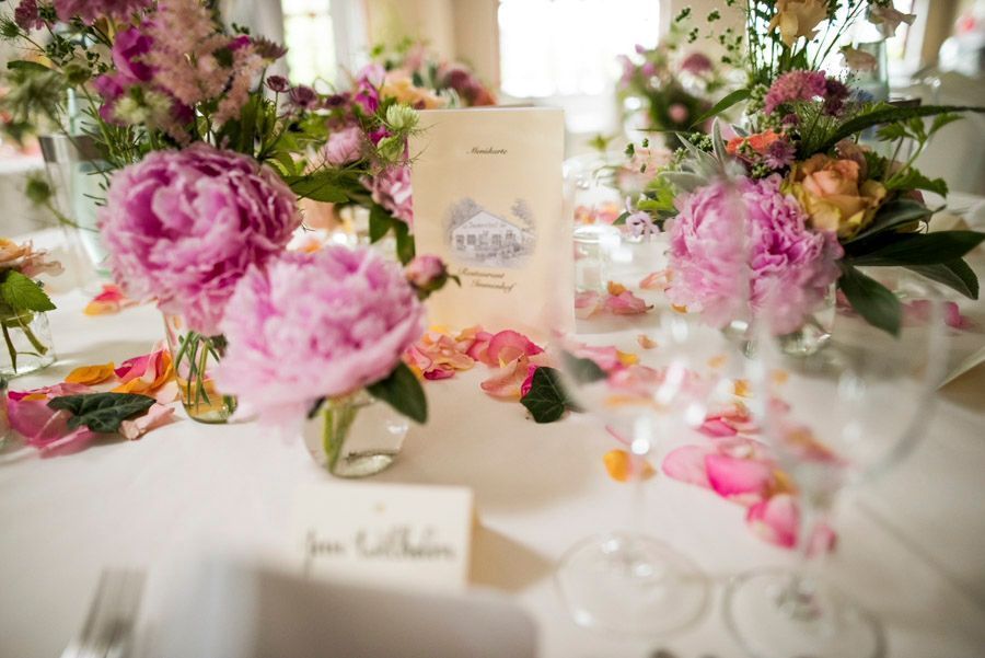 Elegant gedeckter Tisch mit rosa Pfingstrosen, Gläsern und einer cremefarbenen Menükarte in zarten Pastellfarben.