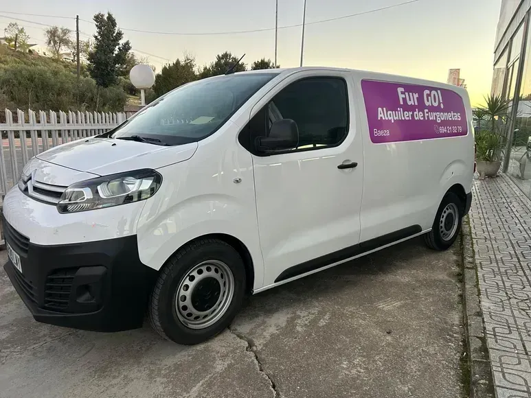 Furgoneta blanca de carga con el logotipo morado de la empresa aparcada en un camino de entrada junto a una valla al atardecer.