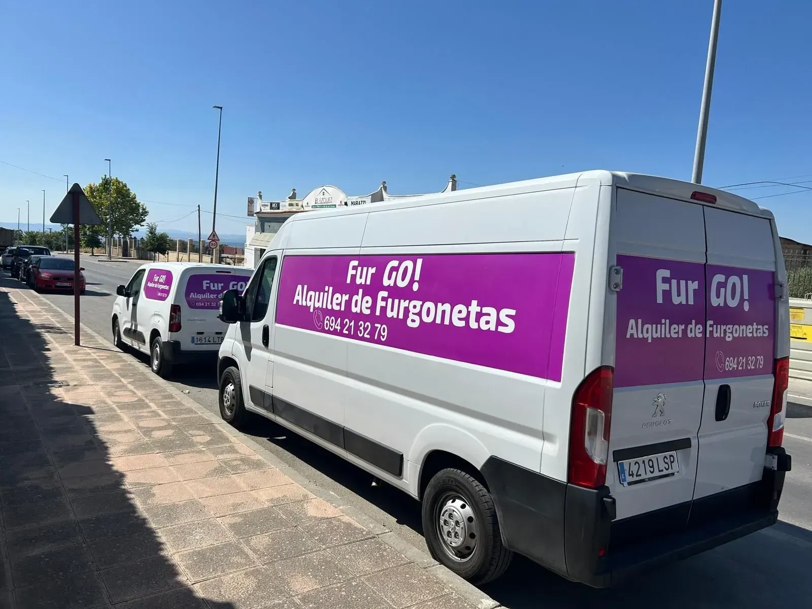 Furgonetas blancas de alquiler con anuncios morados de “Fur GO! Alquiler de Furgonetas” aparcadas en una calle soleada.