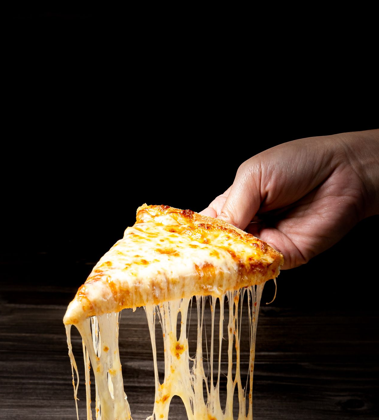 Main tenant une tranche de pizza avec des fils de fromage fondu sur un fond sombre.
