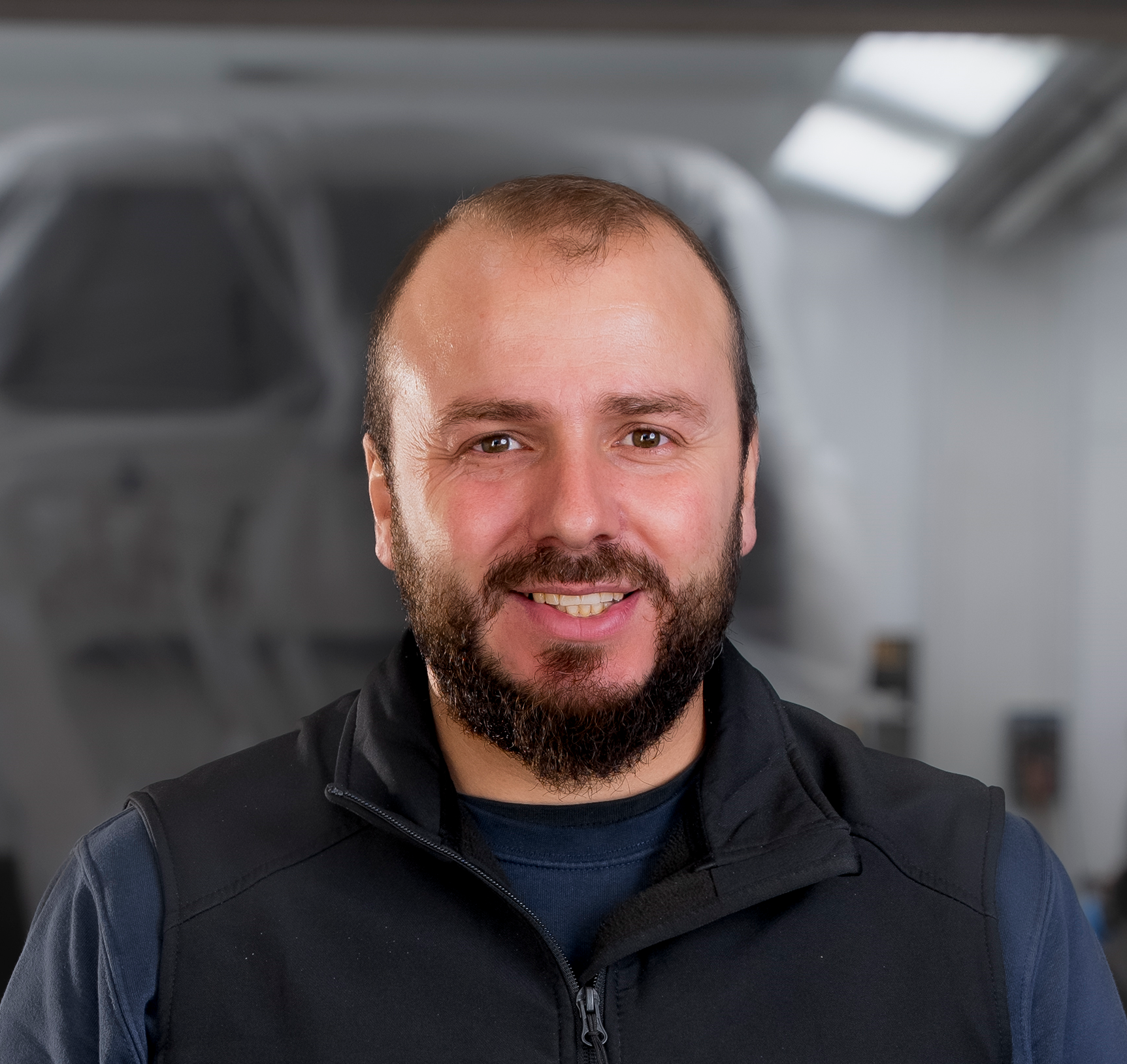 Lächelnder Mann mit Bart und dunklem Hemd. In einer Werkstatt mit dem Logo der Carrosserie Werke AG.