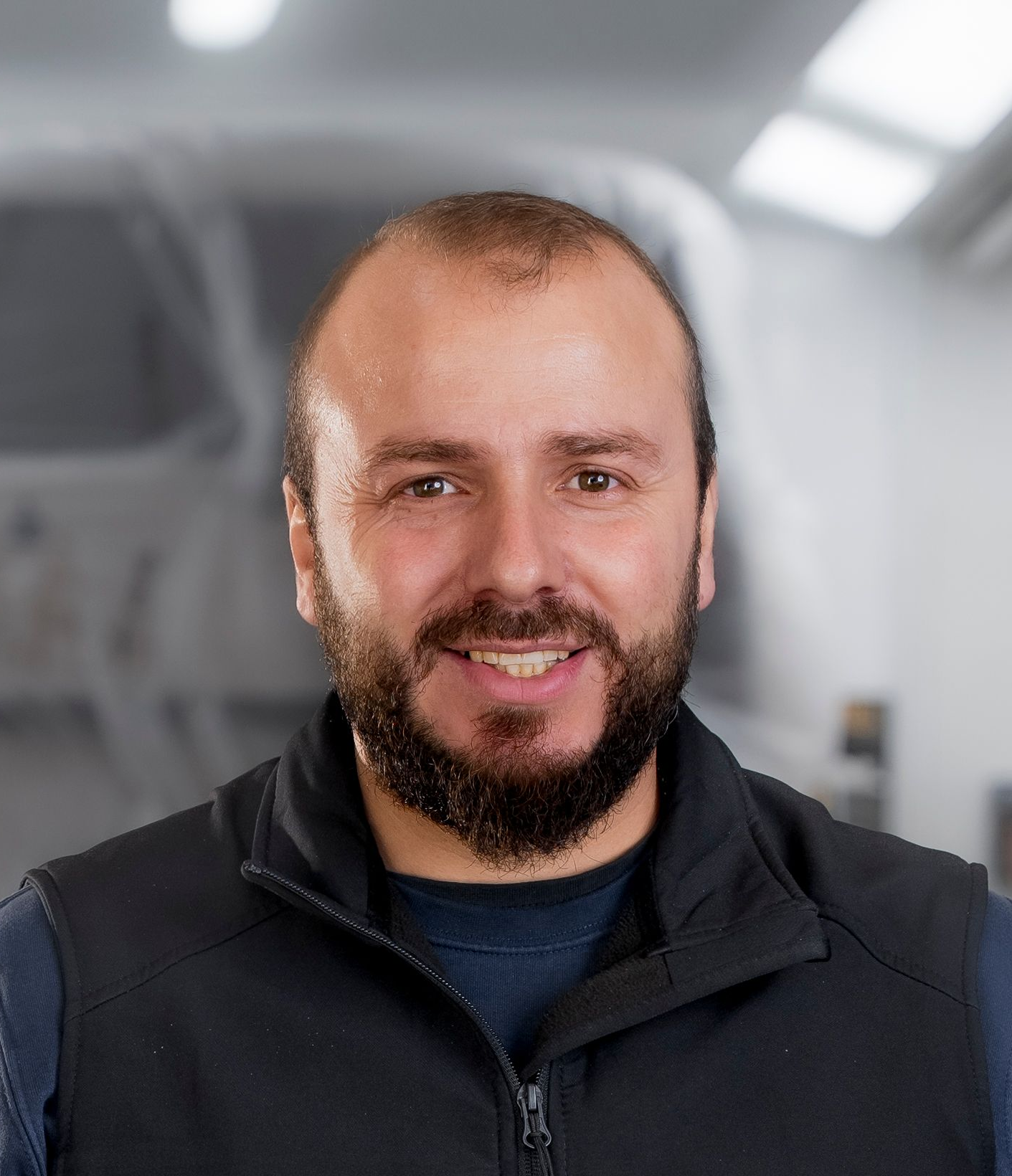 Lächelnder Mann mit Bart und dunklem Hemd. In einer Werkstatt mit dem Logo der Carrosserie Werke AG.