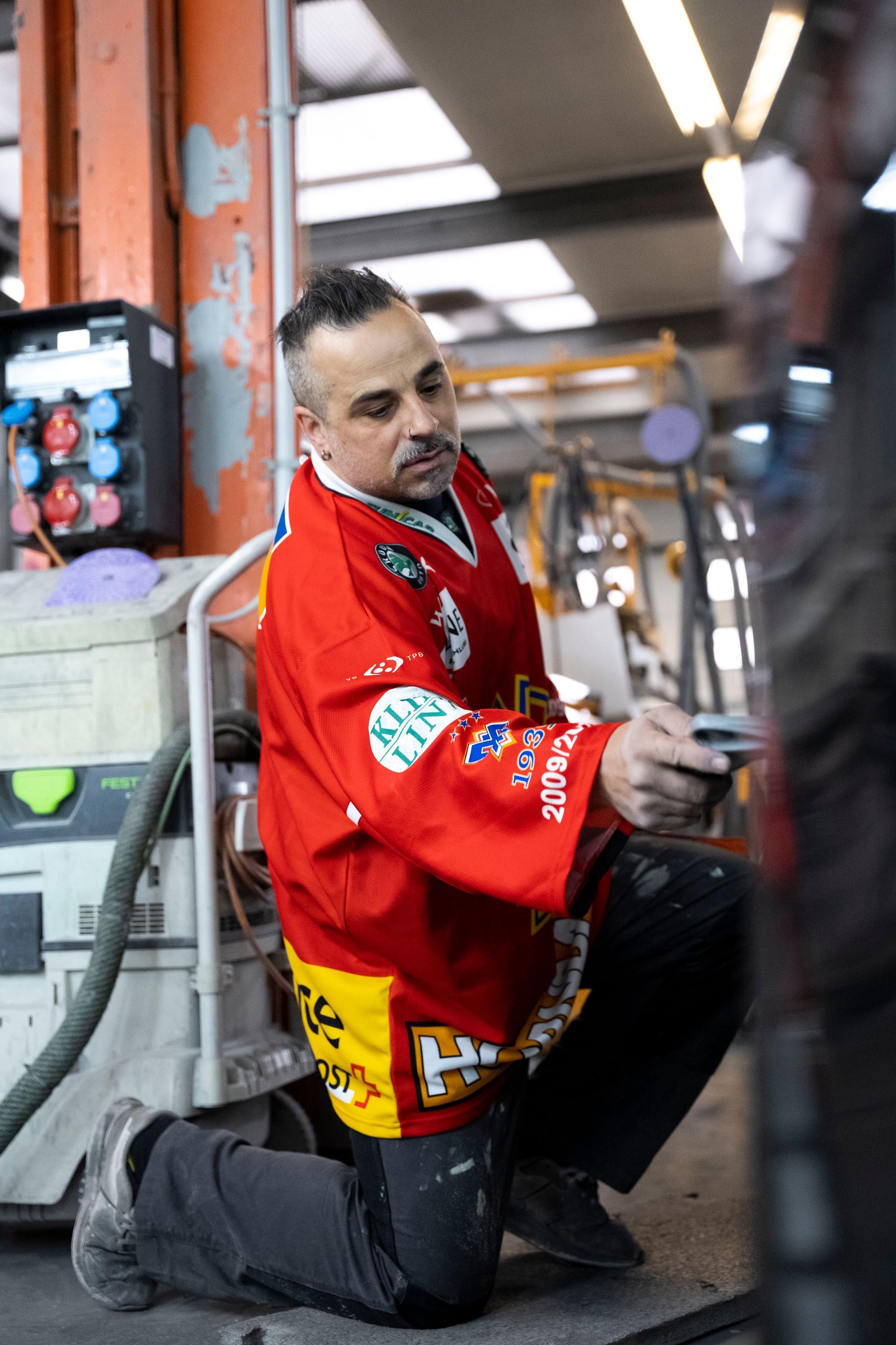 Mann im roten Hockeytrikot kniet und inspiziert etwas. Industrielle Umgebung mit Ausrüstung.