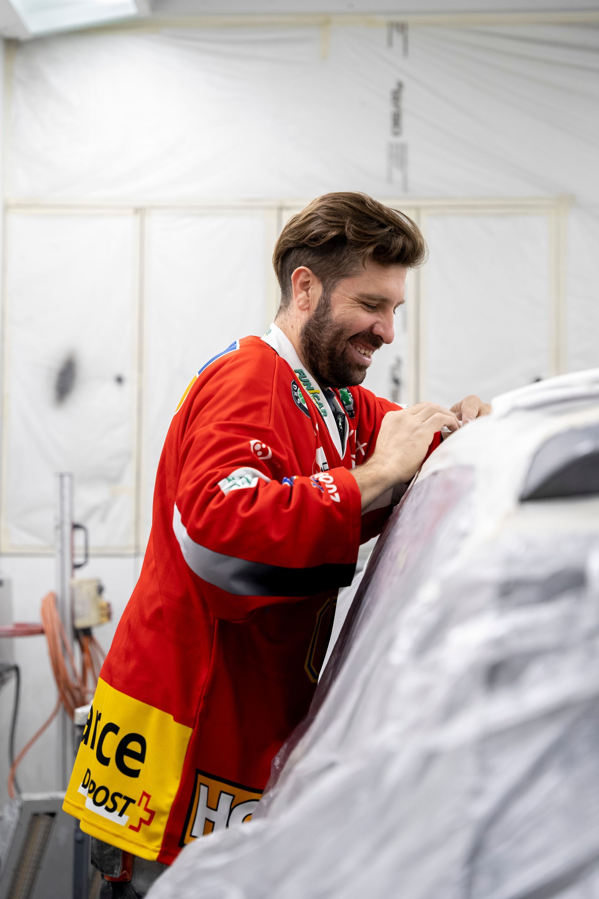 Mann im roten Trikot beugt sich lächelnd über ein Auto in einer Werkstatt.