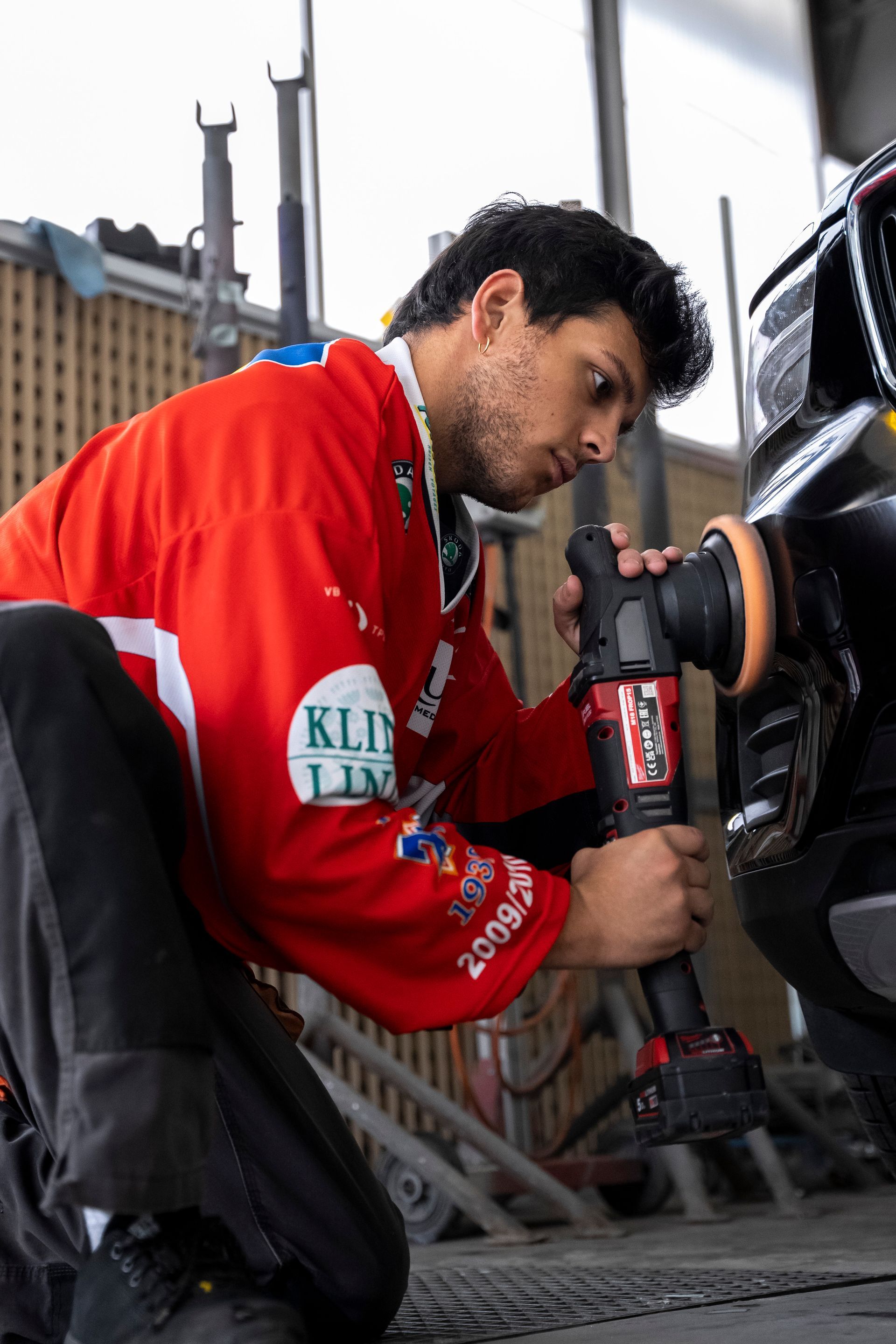Ein Mann im roten Hemd poliert in einer Garage eine schwarze Autostoßstange mit einer elektrischen Poliermaschine.
