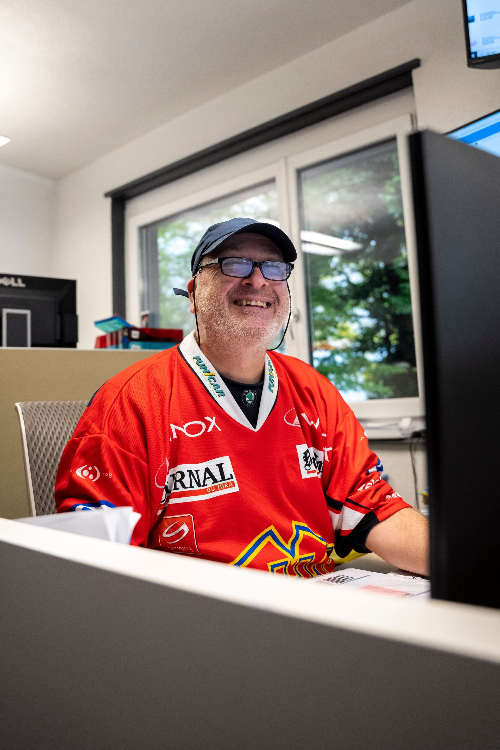 Mann im roten Trikot lächelt, während er mit Hut und Brille an einem Schreibtisch vor einem Computer sitzt.