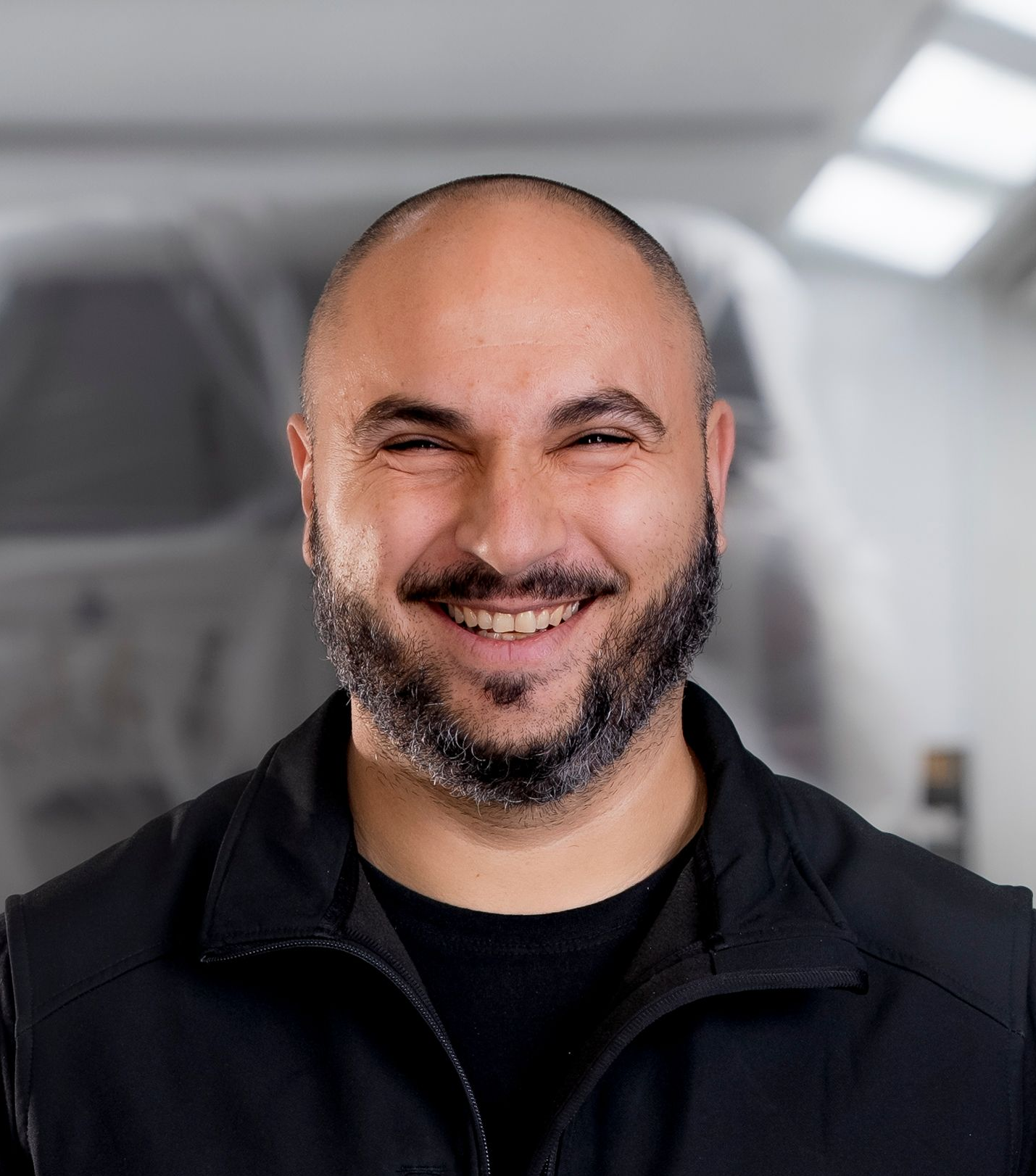 Mann mit ergrauendem Haar und Bart, lächelnd, trägt ein blaues Sweatshirt mit Text, drinnen.