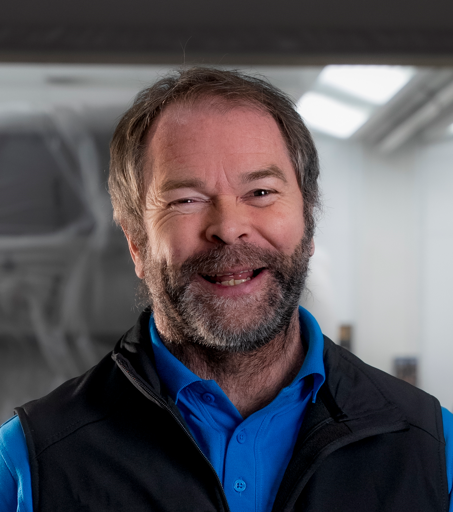 Lächelnder Mann mit dunklem Haar und schwarzer Weste mit Logos in einer Werkstatt.