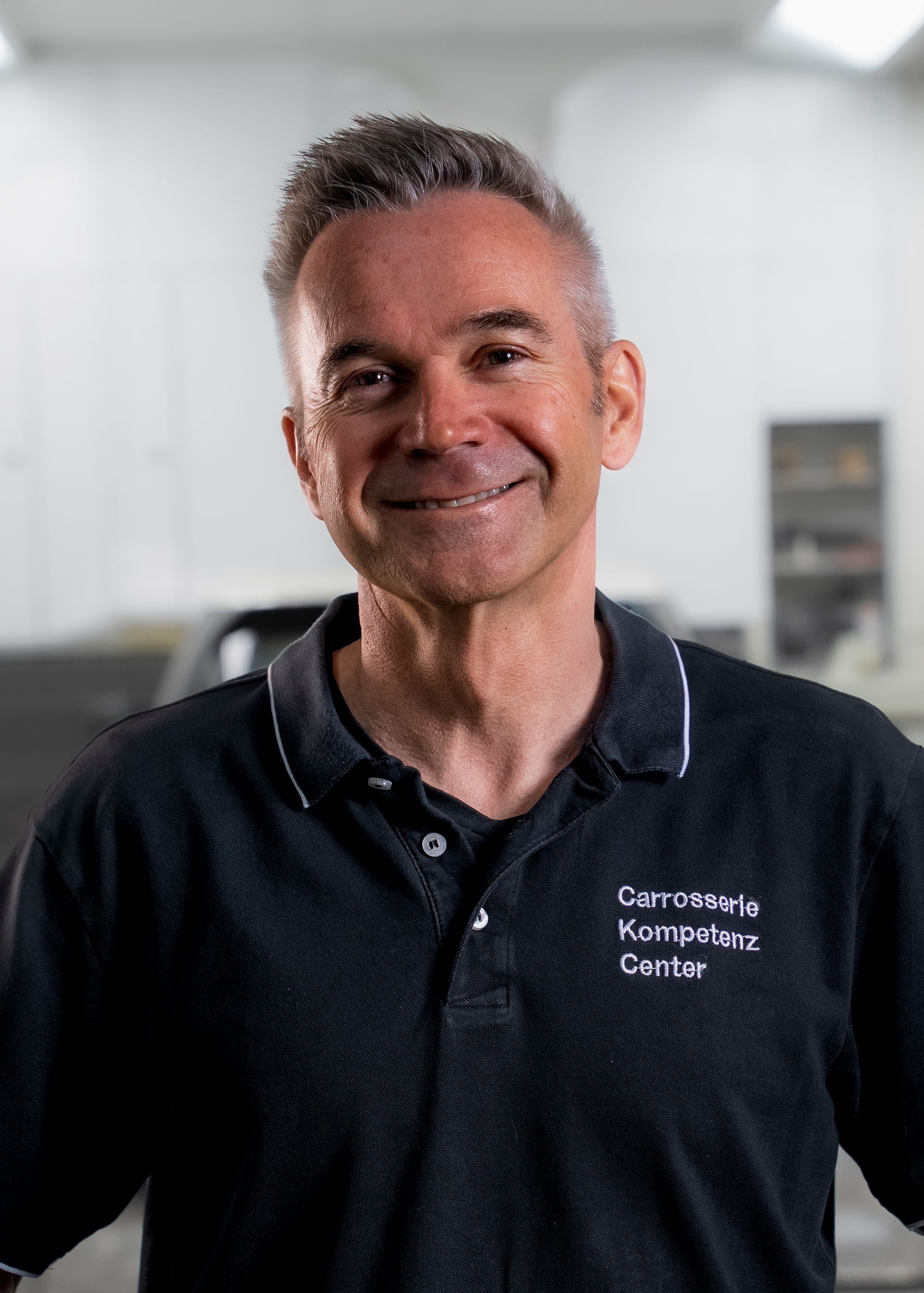 Lächelnder Mann in einer Werkstatt; trägt ein schwarzes Poloshirt mit weißem Kragen und Logo; graues Haar.