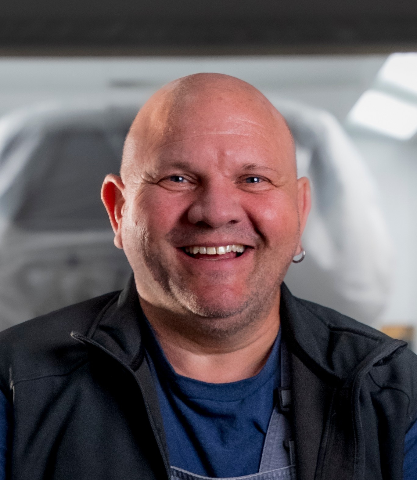 Lächelnder Mann mit dunklem Haar in einem schwarzen Poloshirt, der vor einem verschwommenen Werkstatthintergrund steht.