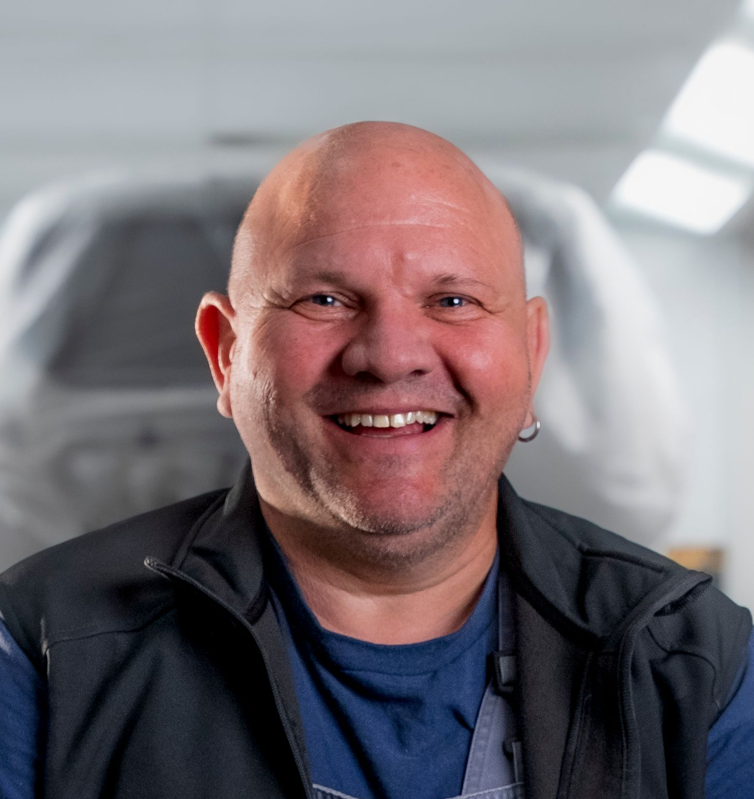 Lächelnder Mann mit dunklem Haar in einem schwarzen Poloshirt, der vor einem verschwommenen Werkstatthintergrund steht.