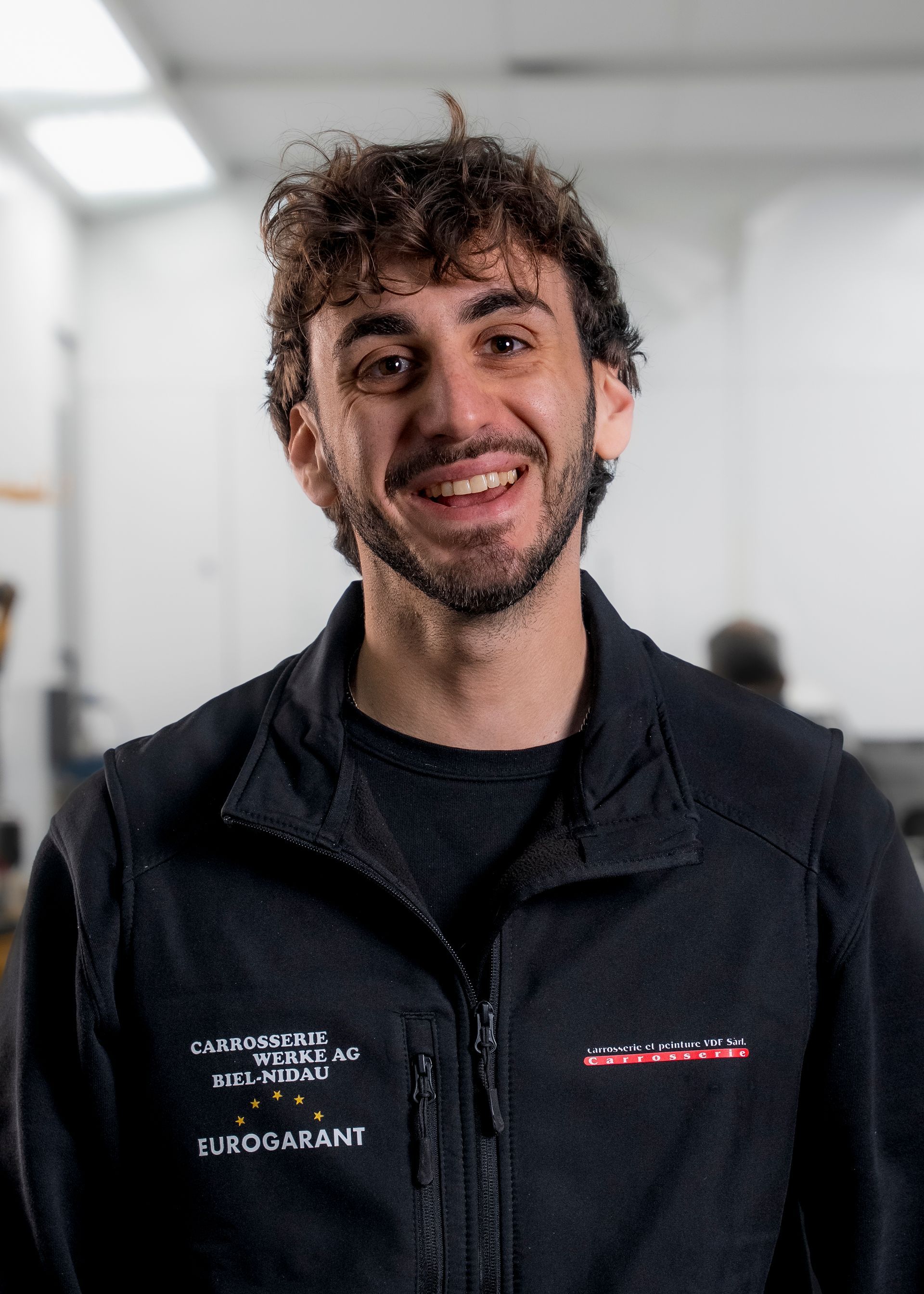 Frau mit kurzen braunen Haaren, lächelnd, mit weißem Blazer und Halskette, drinnen.