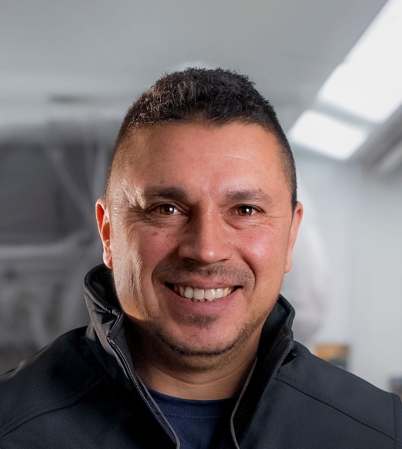 Lächelnder Mann in einer Werkstatt; trägt ein schwarzes Poloshirt mit weißem Kragen und Logo; graues Haar.