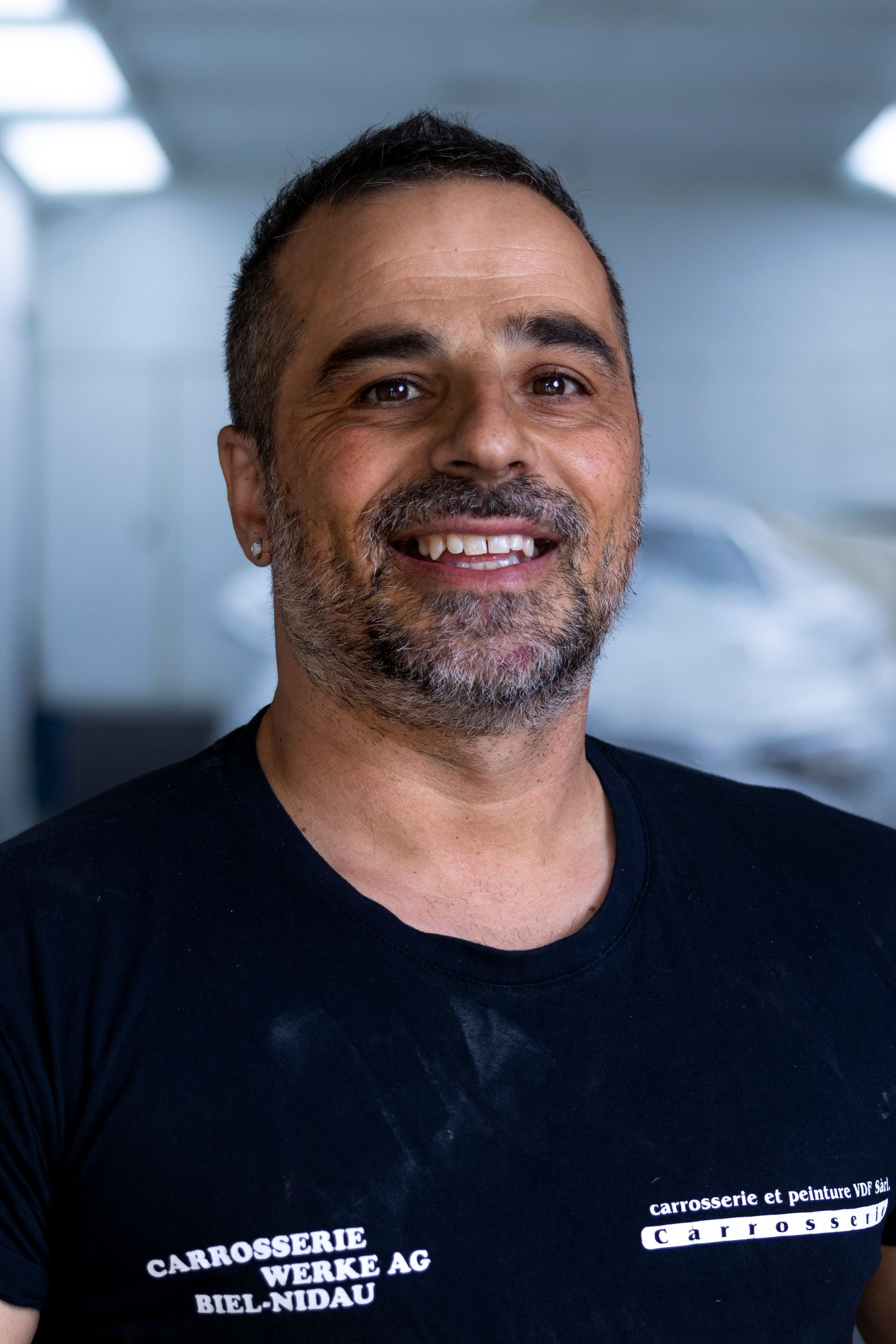 Lächelnder Mann mit dunklem Haar und schwarzer Weste mit Logos in einer Werkstatt.