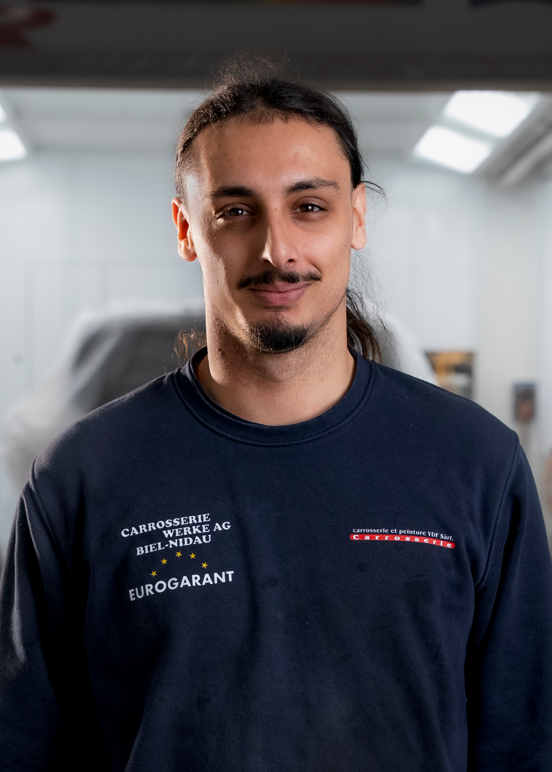 Ein Mann in einem dunkelblauen Hemd lächelt in einer Werkstatt in die Kamera.