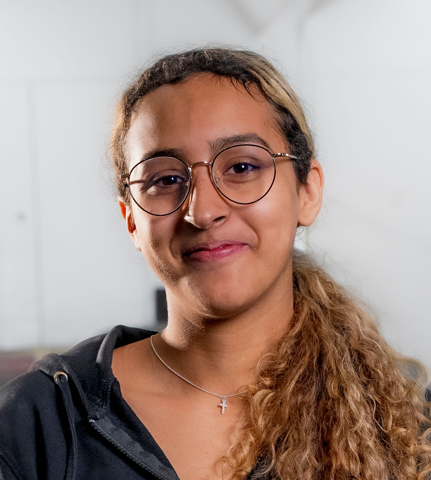 Lächelnder Mann mit dunklem Haar in einem schwarzen Poloshirt, der vor einem verschwommenen Werkstatthintergrund steht.
