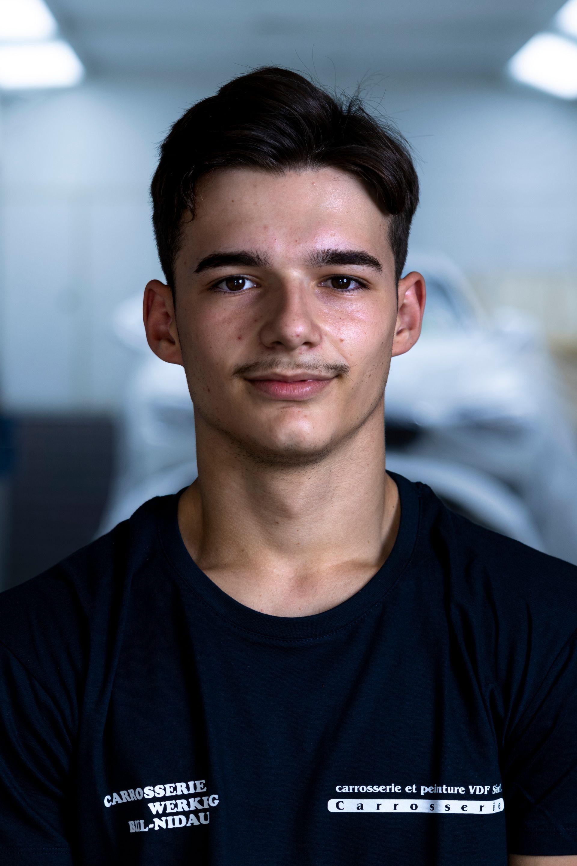 Lächelnder Mann in einer Werkstatt; trägt ein schwarzes Poloshirt mit weißem Kragen und Logo; graues Haar.