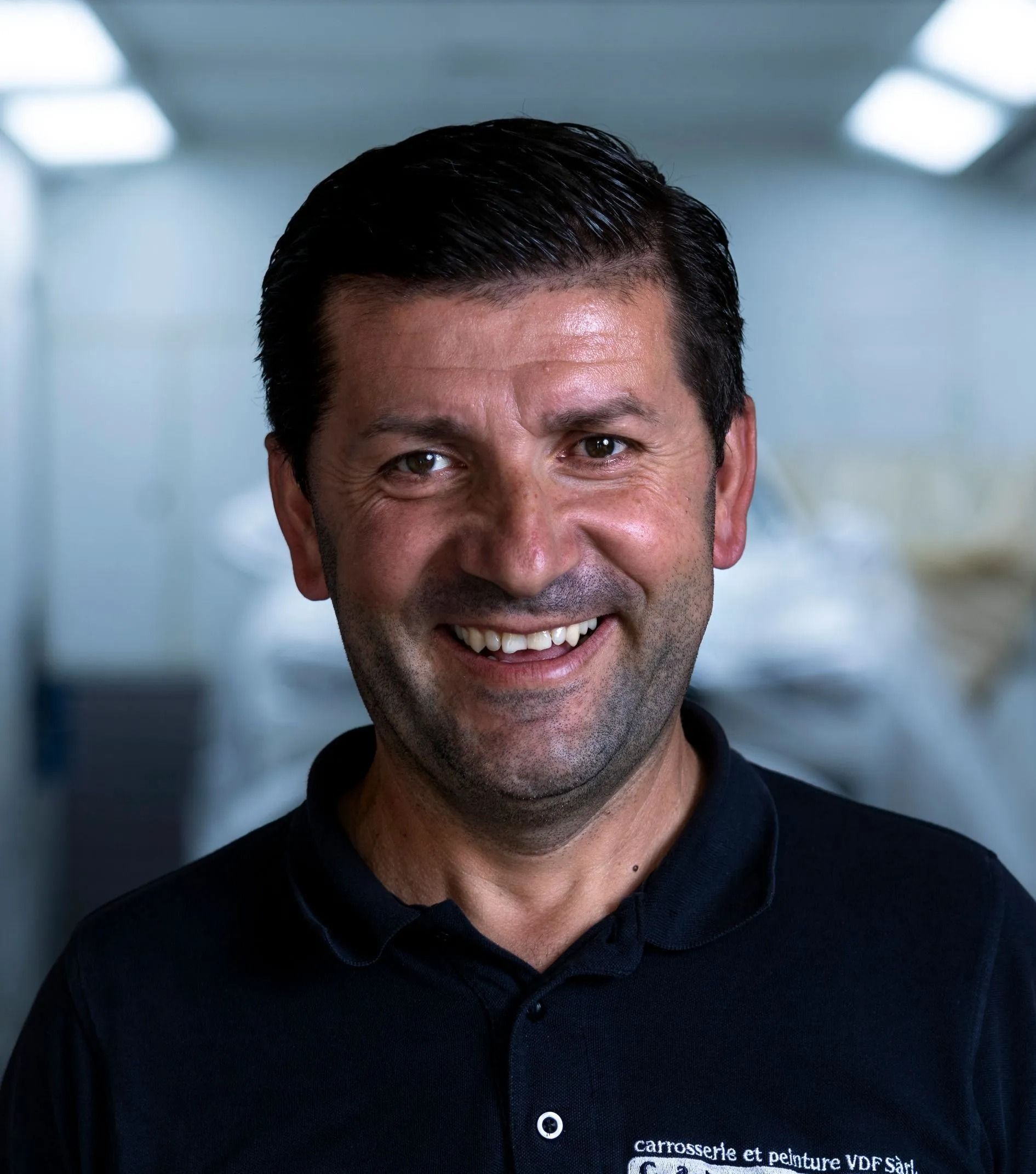 Lächelnder Mann mit Bart und dunklem Hemd. In einer Werkstatt mit dem Logo der Carrosserie Werke AG.