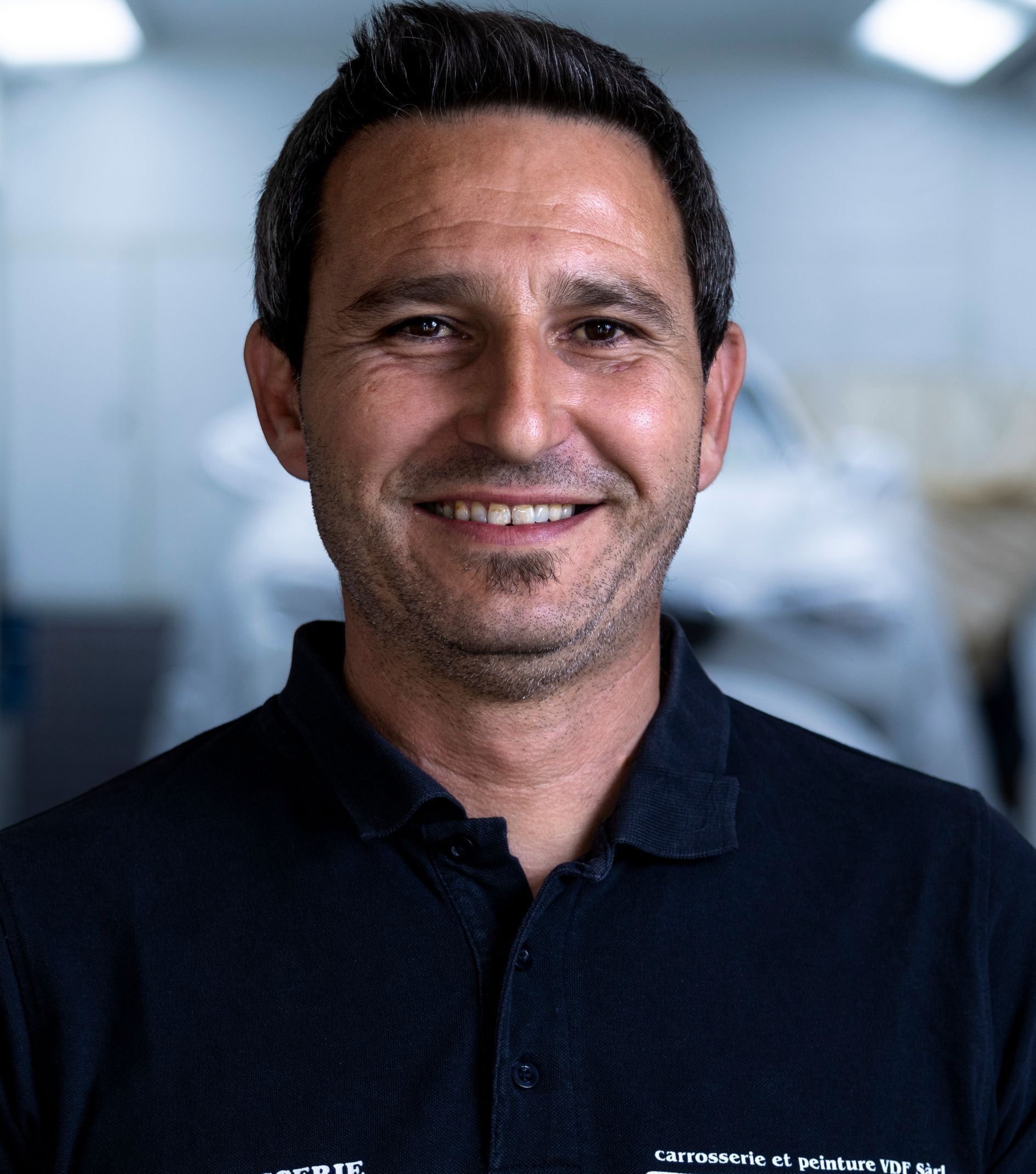 Ein Mann in einem dunkelblauen Hemd lächelt in einer Werkstatt in die Kamera.